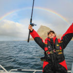 Ein Blinker-Redakteur sitzt samt Angelrute und Köder auf einem Boot in Norwegen. I Hintergrund ist ein Regenbogen zu sehen.