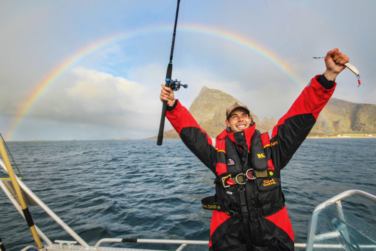 Ein Blinker-Redakteur sitzt samt Angelrute und Köder auf einem Boot in Norwegen. I Hintergrund ist ein Regenbogen zu sehen.