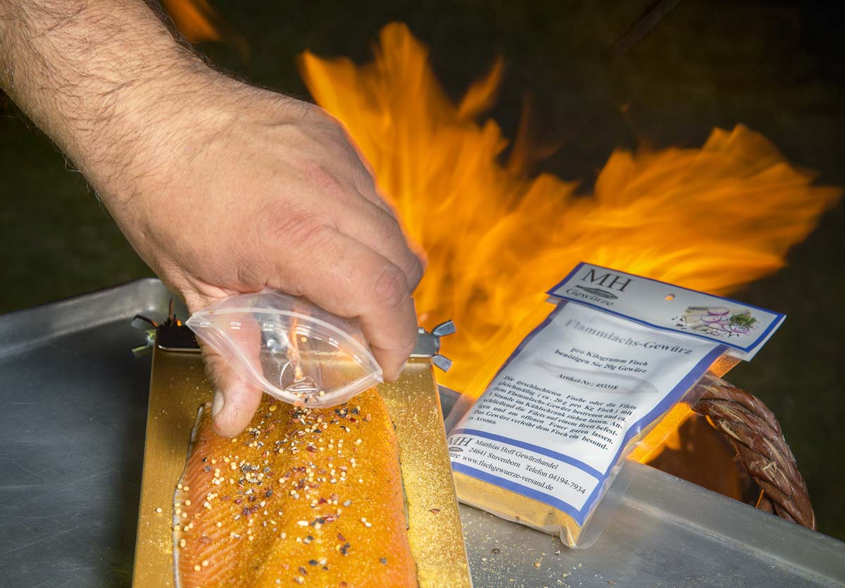 Zuerst wird das Filet mit der Hautseite auf das Brett gelegt und mit Flammlachs-Gewürz bestreut. Danach gibt man noch einige ganze Pfefferkörner auf das Filet. Diese geben dem Fisch die besondere Note.