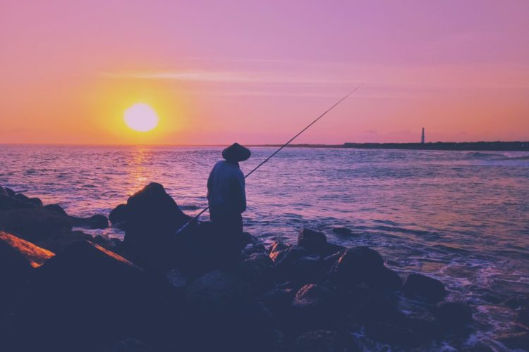 Ein angler steht mit Rute am Wasser und beobachtet den Sonnenuntergang