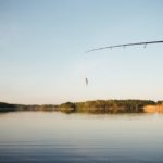 Ein angler steht am Wasser, zu sehen sind Wasser, Bäume, die Rute und ein kleiner Köder.