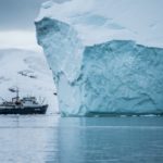 Auf dem Wasser fährt ein kleines Schiff hinter einen großen Eisberg.