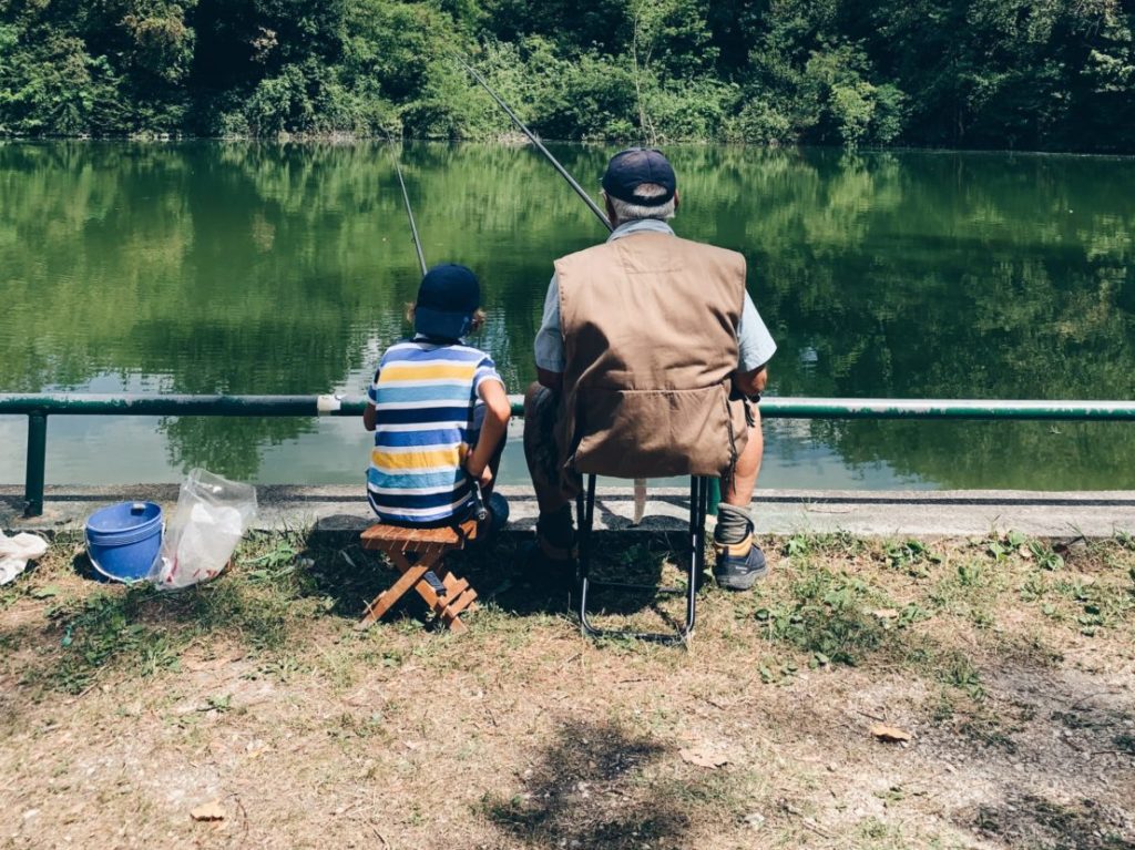 Ein Junge und ein älterer Mann sitze an einem Ufer mit ihren Angeln im Wasser.