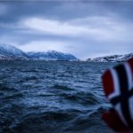 Im Vordergrund eine unscharfe norwegische Flagge aus dem Meer. Im Hintergrund Meer und am Horizont die verschneiten Berge und Lichter einer Stadt.