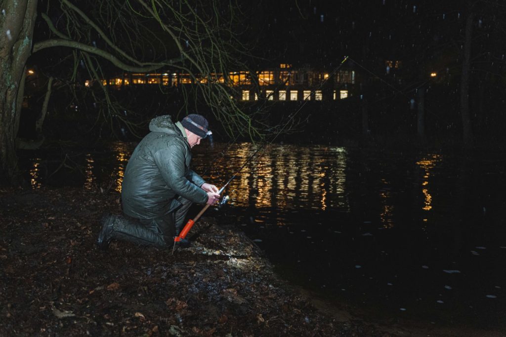 Ein Angler kniet im Winter an einem kleinen Fluss. Er hat gerade ausgeworfen und spannt nun die Schnur.