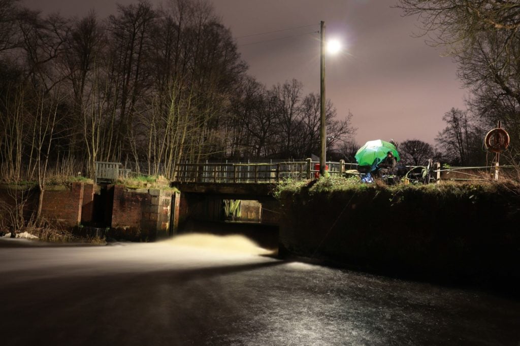 Ein Wehr im November, unter einem grünen Schirm sitzt ein Angler