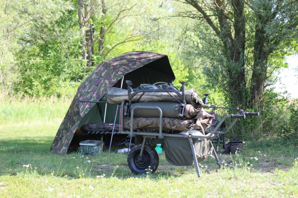 Karpfenzelt im späten Frühling vor grünen Büschen, davor ein bepackter Trolley