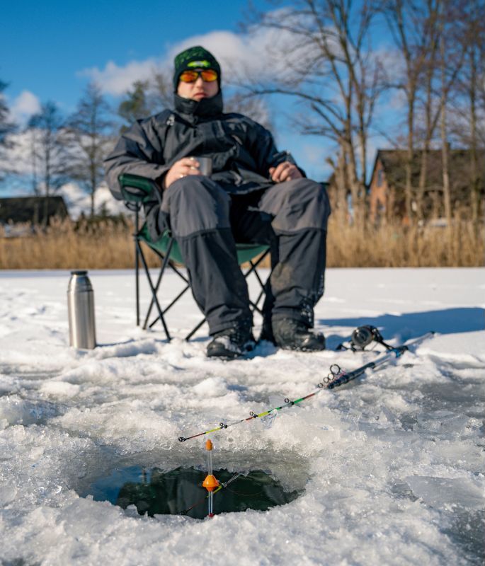 Angler sitzt beim Eisangeln auf Hecht neben dem Eisloch