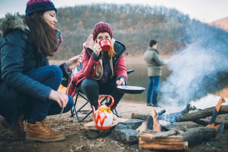 Warme Getränke machen warme Füße: Zwei Frauen am Feuer, ein Mann im Hintergrund angelt